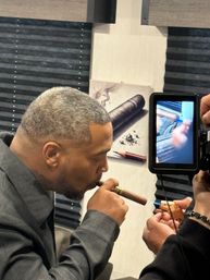 Profile of a man lighting a cigar indoors while another hand holds a lighter and a camera monitor records, lounge-style backdrop with cigar artwork.