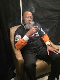 Bearded man relaxing in a leather armchair in a dim indoor lounge, exhaling cigar smoke while wearing a black shirt with orange sleeves and a gold watch.