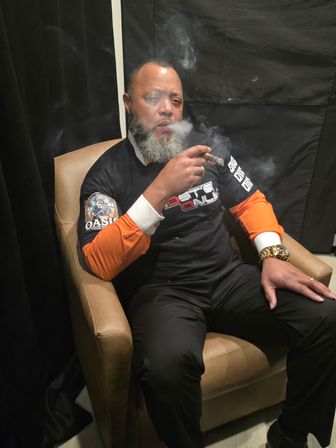 Bearded man relaxing in a leather armchair in a dim indoor lounge, exhaling cigar smoke while wearing a black shirt with orange sleeves and a gold watch.