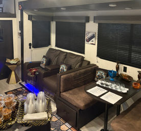 Modern RV interior lounge in a travel trailer with brown leather reclining seats, bench-style dinette set with glasses and snacks, dark pleated window shades and blue accent lighting.