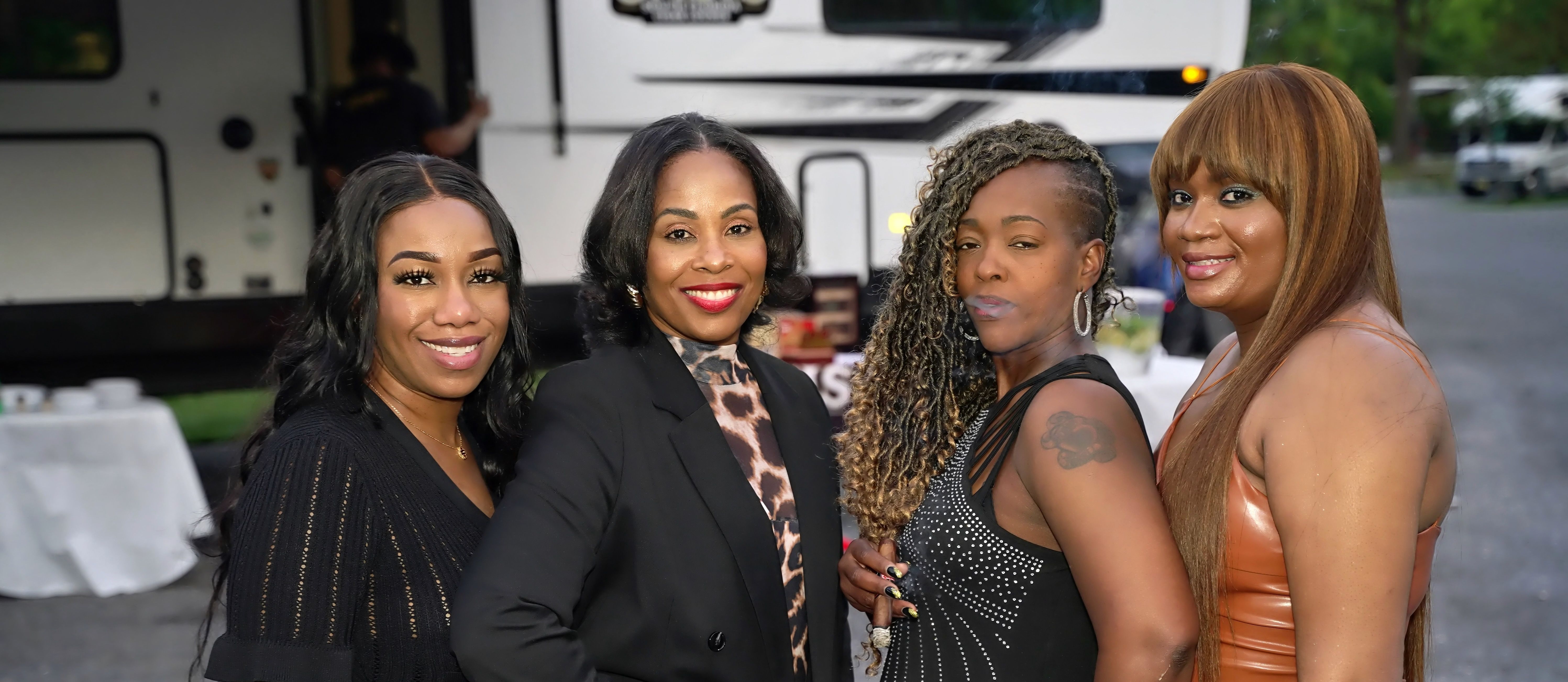 Four women dressed up and smiling pose together in front of a parked RV at an outdoor gathering, featuring a black blazer with leopard-print blouse, a studded black dress, a shimmery leather dress, and one with braided side-shave and a shoulder tattoo.