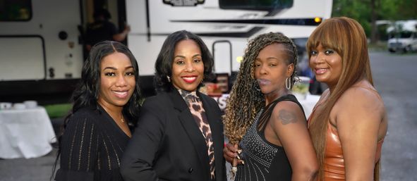 Four women dressed up and smiling pose together in front of a parked RV at an outdoor gathering, featuring a black blazer with leopard-print blouse, a studded black dress, a shimmery leather dress, and one with braided side-shave and a shoulder tattoo.