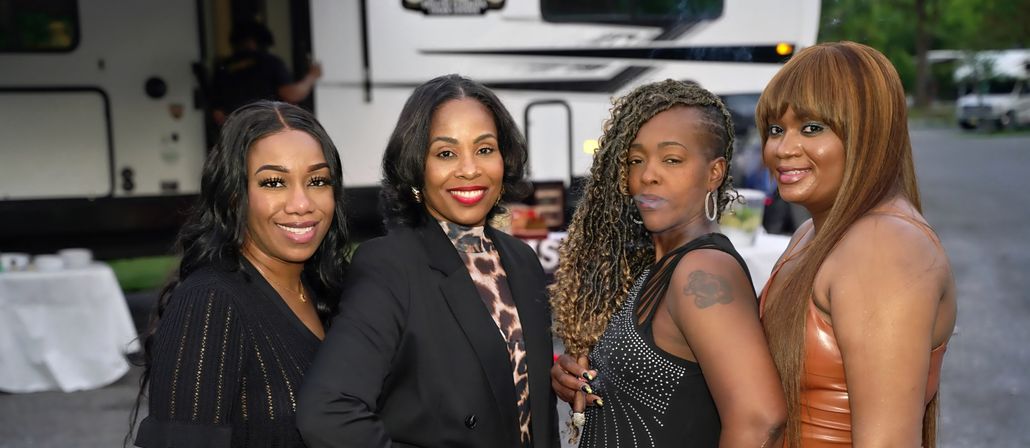 Four women dressed up and smiling pose together in front of a parked RV at an outdoor gathering, featuring a black blazer with leopard-print blouse, a studded black dress, a shimmery leather dress, and one with braided side-shave and a shoulder tattoo.