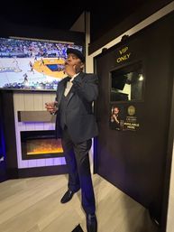 Well-dressed man in a dark suit and cap enjoying a cigar and drink inside a cozy lounge by a 'VIP ONLY' door, with a basketball game on the wall TV and an electric fireplace below.