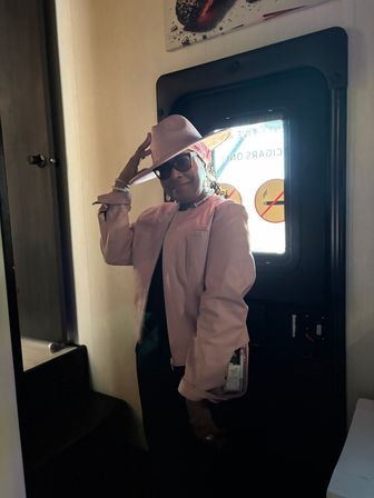 Stylish person in a pink wide-brim hat and matching jacket with sunglasses, striking a playful pose indoors beside a backlit door/window displaying no-smoking signs, holding a clear handbag.