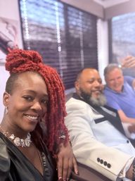 Three friends posing for a selfie at an indoor celebration: smiling woman with bright red dreadlocks and a statement necklace in the foreground, a bearded man in a white tux jacket beside her, and a man in a blue shirt raising a glass in the background near window blinds.