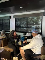 Two people talking at a round camper dinette inside an RV interior, drink and blue bowl on the table, dark pleated window shades and booth seating with another person seated in the background.
