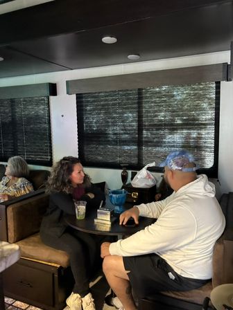 Two people talking at a round camper dinette inside an RV interior, drink and blue bowl on the table, dark pleated window shades and booth seating with another person seated in the background.
