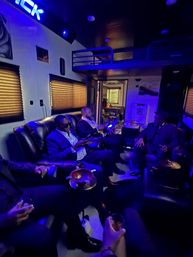 Blue-lit luxury mobile lounge with men in suits relaxing on leather couches, holding drinks and cigars around ashtray-topped tables, lofted bunk and window blinds lending an intimate urban nightlife vibe.