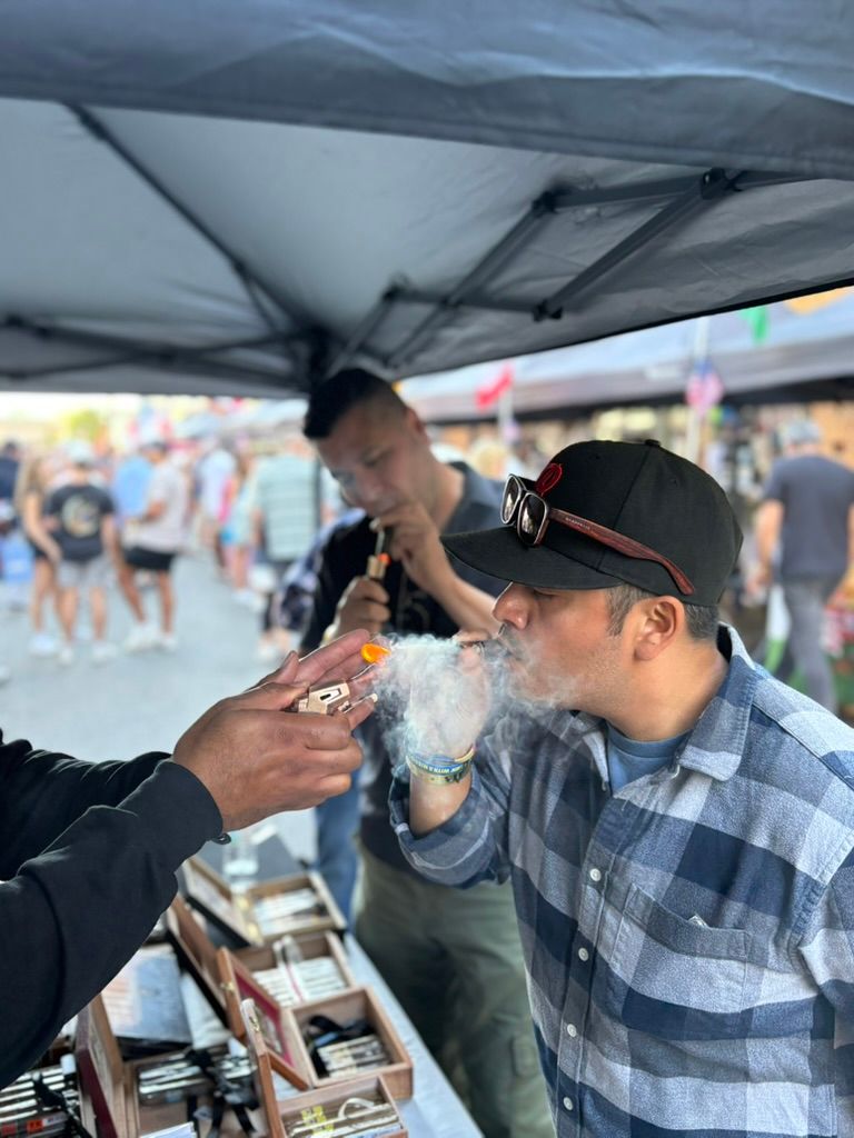 Man in plaid shirt and baseball cap exhales a cloud of vapor while a vendor lights his cigar at a lively outdoor street market stall under a canopy, with boxes of cigars and a blurred crowd in the background.