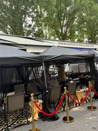 Stylish outdoor cigar lounge pop-up on a city street with black canopy tents, mesh curtains, seating, patterned rug and gold stanchions with red velvet ropes.