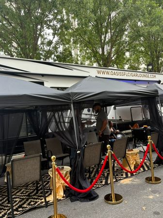 Stylish outdoor cigar lounge pop-up on a city street with black canopy tents, mesh curtains, seating, patterned rug and gold stanchions with red velvet ropes.