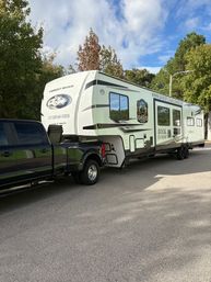 Sleek black pickup truck hauling a large white fifth-wheel RV, parked on a tree-lined suburban street under a bright blue sky.