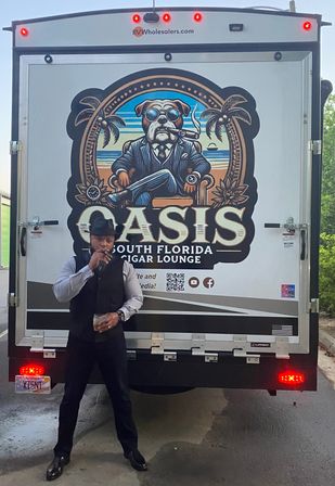 Dapper man in a fedora smoking a cigar and holding a drink in front of a delivery truck with a bold mural of a cigar-smoking bulldog, palm trees and 'South Florida cigar lounge' text.