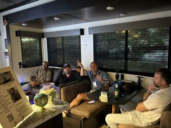 Four men relaxing in a cozy RV interior lounge with dark window shades, small table with drinks and a phone, a jar of cash and a decorative globe on the counter — casual evening hangout.