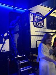 Nighttime scene of a mobile cigar lounge trailer lit by blue LED lights, fold-out stairs with velvet ropes, a person stepping out of the doorway and a blurred guest in a white dress.