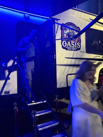 Nighttime scene of a mobile cigar lounge trailer lit by blue LED lights, fold-out stairs with velvet ropes, a person stepping out of the doorway and a blurred guest in a white dress.