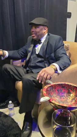 Seated person in a tuxedo and flat cap relaxing in a leather armchair at a formal event or cocktail lounge, bow tie and vest visible, black curtain backdrop, decorative red bowl and wine glass on side table, water bottle on floor.