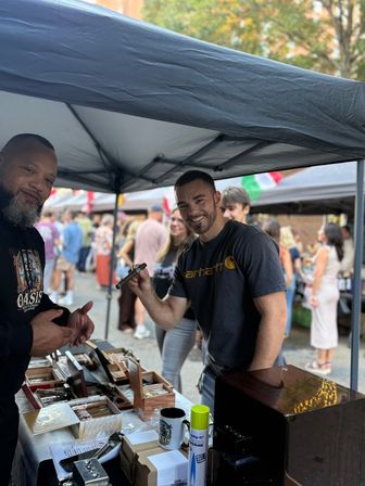 Smiling man holds a cigar at a vendor tent with wooden cigar boxes and accessories on the table, bustling outdoor street market crowd in the background.