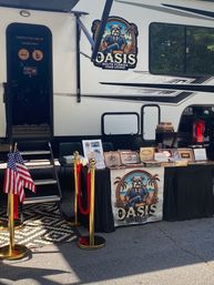 Mobile cigar lounge setup beside a white RV at an outdoor Florida event with a table of cigar boxes, red velvet ropes and American flags