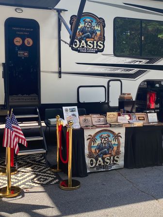 Mobile cigar lounge setup beside a white RV at an outdoor Florida event with a table of cigar boxes, red velvet ropes and American flags