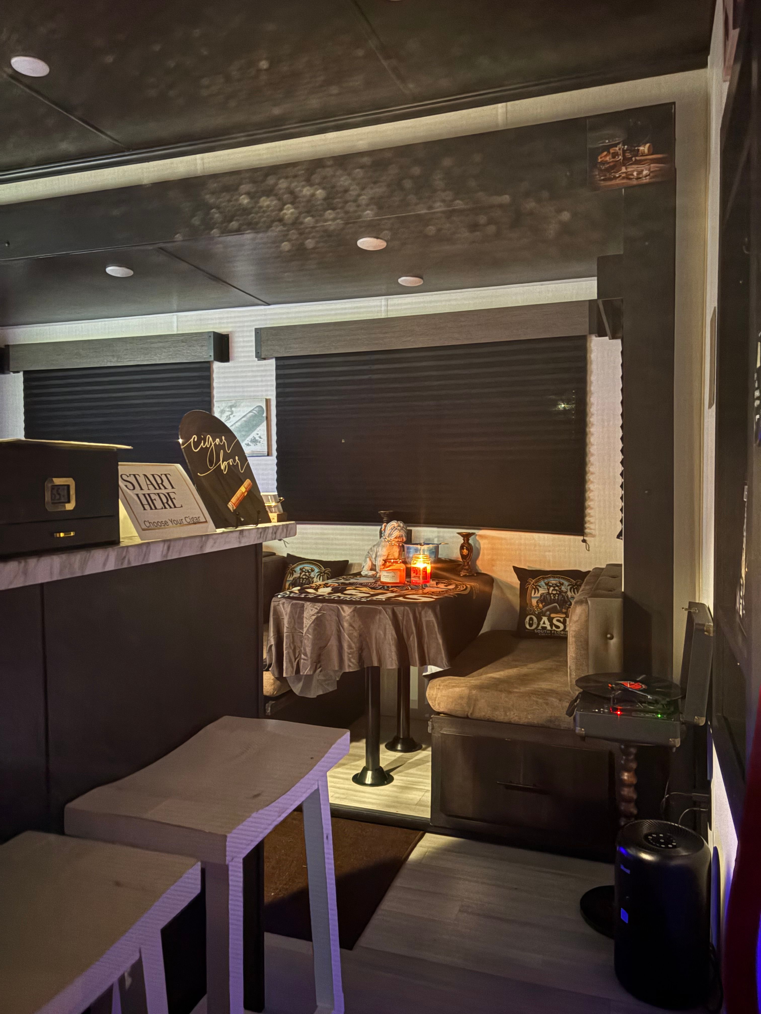 Cozy candlelit RV dining nook with round table, plush bench seating, small bar counter and two stools under dark window shades.