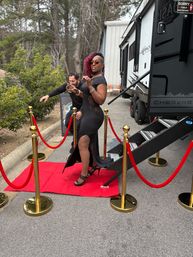 Woman in black evening gown and heels striking a pose on a red carpet with gold stanchions and red ropes next to a travel trailer in an outdoor wooded parking area.