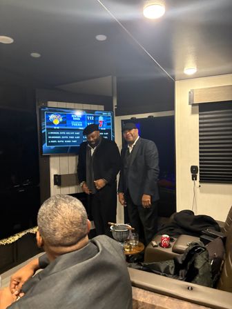 Two men in suits smiling in a cozy indoor lounge watching New York Knicks vs Philadelphia 76ers on a wall TV, drinks on a small table
