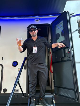 Man in black athletic wear and sunglasses standing on the steps of a camper RV, holding a plastic cup of beer under blue LED awning lights, smiling at the camera.