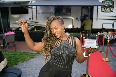 Smiling woman with braided hair on a red carpet at an outdoor cigar-lounge event, holding a lit cigar in one hand and a small award trophy in the other with an RV trailer backdrop.
