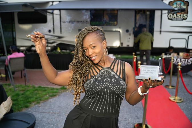 Smiling woman with braided hair on a red carpet at an outdoor cigar-lounge event, holding a lit cigar in one hand and a small award trophy in the other with an RV trailer backdrop.