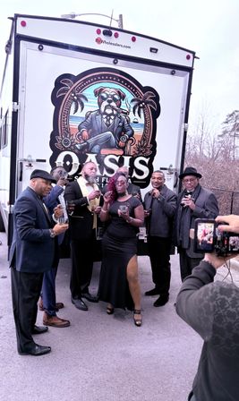 Six adults dressed up posing in an outdoor parking-lot event in front of a colorful trailer mural, smoking cigars and holding whiskey glasses while a photographer films with a phone.
