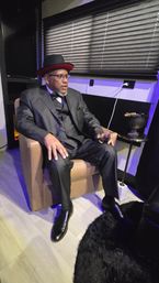 Man in a three-piece suit, bow tie and black hat with red brim seated in a leather armchair holding a glass of whiskey, cigar resting on a side table in a dim modern lounge