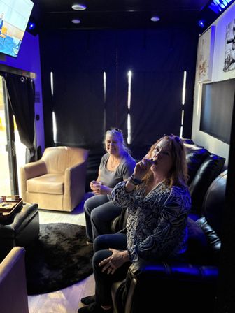 Two women relaxing in a cozy, blue-lit indoor lounge with leather recliners and a plush rug — one woman takes a puff from a vape while the other smiles near an armchair and wall-mounted TV.
