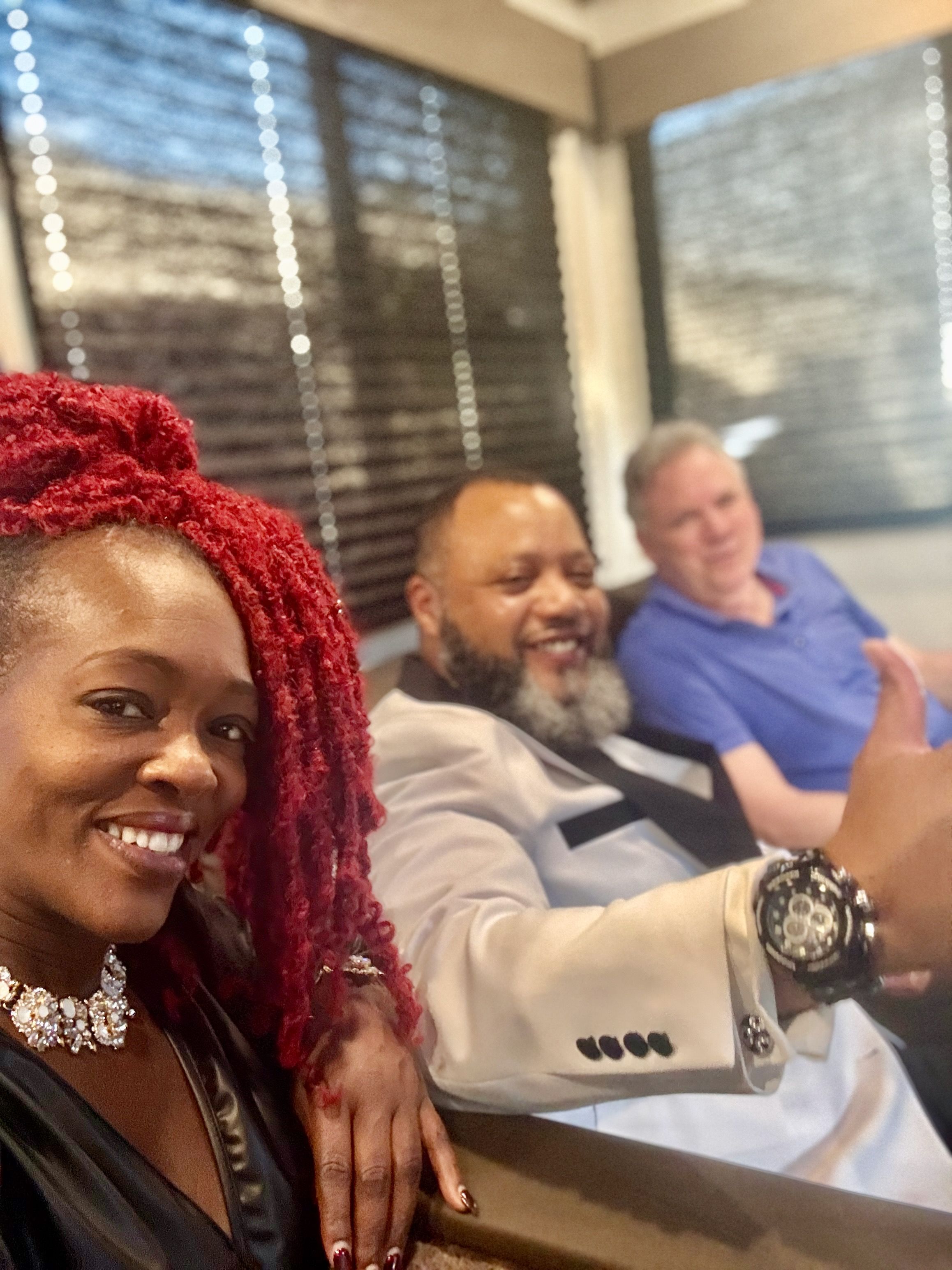 Three friends smiling in a cozy indoor lounge — woman with bright red braided hair and a statement necklace takes a selfie, a bearded man in a light blazer gives a thumbs-up showing a bold wristwatch, and a man in a blue polo relaxes on a sofa with window blinds behind them.