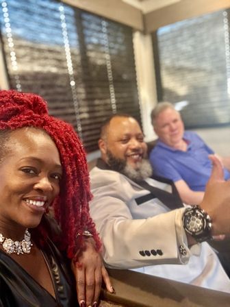 Three friends smiling in a cozy indoor lounge — woman with bright red braided hair and a statement necklace takes a selfie, a bearded man in a light blazer gives a thumbs-up showing a bold wristwatch, and a man in a blue polo relaxes on a sofa with window blinds behind them.