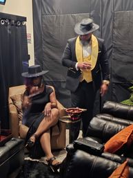 Two elegantly dressed people in a dim lounge with a smoky speakeasy vibe — a seated woman in a black dress and wide-brim hat crossing her legs, and a standing man in a black suit with a gold vest and fedora holding a drink beside a small table with a cigar bowl and leather recliners.