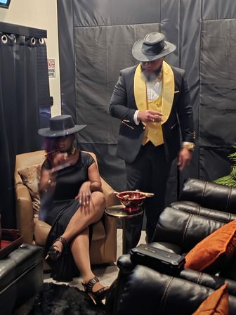 Two elegantly dressed people in a dim lounge with a smoky speakeasy vibe — a seated woman in a black dress and wide-brim hat crossing her legs, and a standing man in a black suit with a gold vest and fedora holding a drink beside a small table with a cigar bowl and leather recliners.