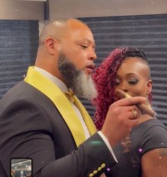 Two stylish guests at a formal indoor celebration: man in a black tuxedo with gold lapel and bow tie holding a cigar, woman with red curly hair and shaved side smiling behind him.