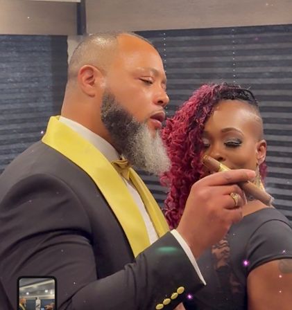 Two stylish guests at a formal indoor celebration: man in a black tuxedo with gold lapel and bow tie holding a cigar, woman with red curly hair and shaved side smiling behind him.