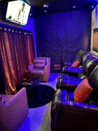 Cozy home theater/media room with black leather recliners and orange throw pillows, beige armchairs, ottoman and round black rug, blue LED ambient lighting, wall-mounted TV and heavy curtains.