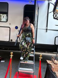 Woman with purple hair stepping down a metal ladder from a trailer onto a red carpet at an outdoor street event, wearing a black tank, tropical-print wide-leg pants, white sneakers, layered necklaces, crossbody bag and sunglasses on her head.