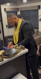 Bearded man in a black suit with a bright yellow lapel leaning over a marble countertop in a cozy backstage dressing room, arranging a gold tray with a tablet and small decorative airplane under warm overhead lights.