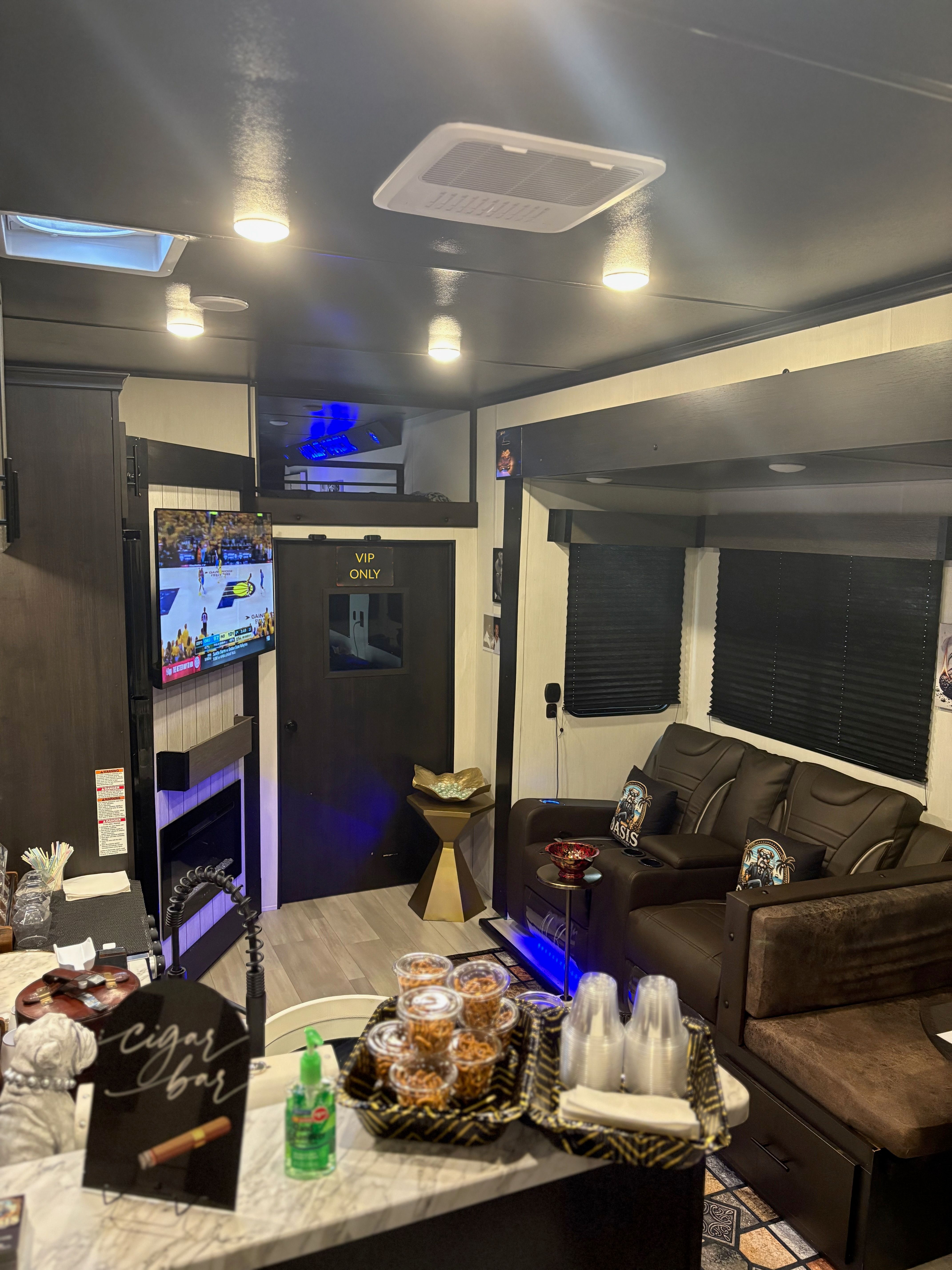 Cozy luxury RV lounge interior with leather reclining theater seats, wall-mounted TV showing a basketball game, a 'VIP ONLY' door, marble countertop with snack cups and disposable cups, and blue accent lighting.