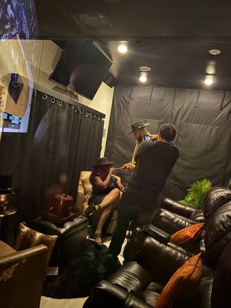 Dimly lit cozy lounge interior with leather recliners and plush pillows; a seated woman in a black dress and hat chats with two men—one showing a phone—against black curtains and warm ceiling lights