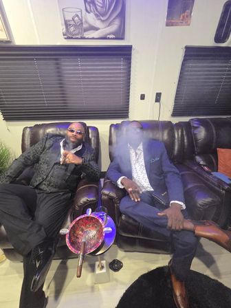 Two men in suits relaxing on dark leather recliners in a cozy indoor cigar lounge, one exhaling smoke with cigars and a colorful ashtray table in front, black window blinds and wall art behind them.