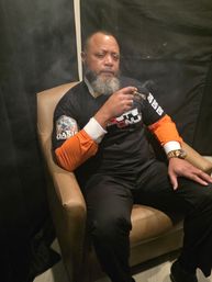 Bearded man in a black-and-orange shirt seated in a tan armchair, holding a lit cigar with smoke curling around him and wearing a gold watch — indoor cigar lounge vibe.