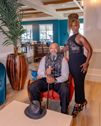 Stylish hotel lounge portrait of a seated man in a patterned vest holding a cane with a fedora on the table and a woman in an elegant black dress standing beside him, teal-accented room with potted palm and wooden floors.