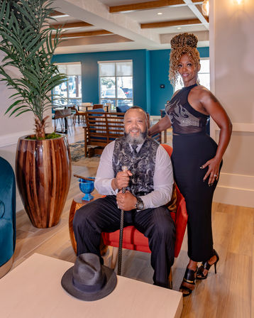 Stylish hotel lounge portrait of a seated man in a patterned vest holding a cane with a fedora on the table and a woman in an elegant black dress standing beside him, teal-accented room with potted palm and wooden floors.