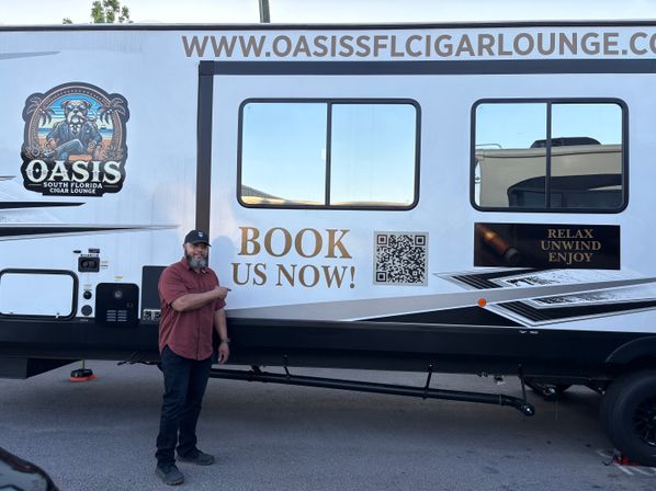 Person in a cap points at the side of a white mobile cigar lounge trailer parked outdoors, featuring two windows, a QR code and bold 'BOOK US NOW!' lettering with a 'RELAX UNWIND ENJOY' sign.